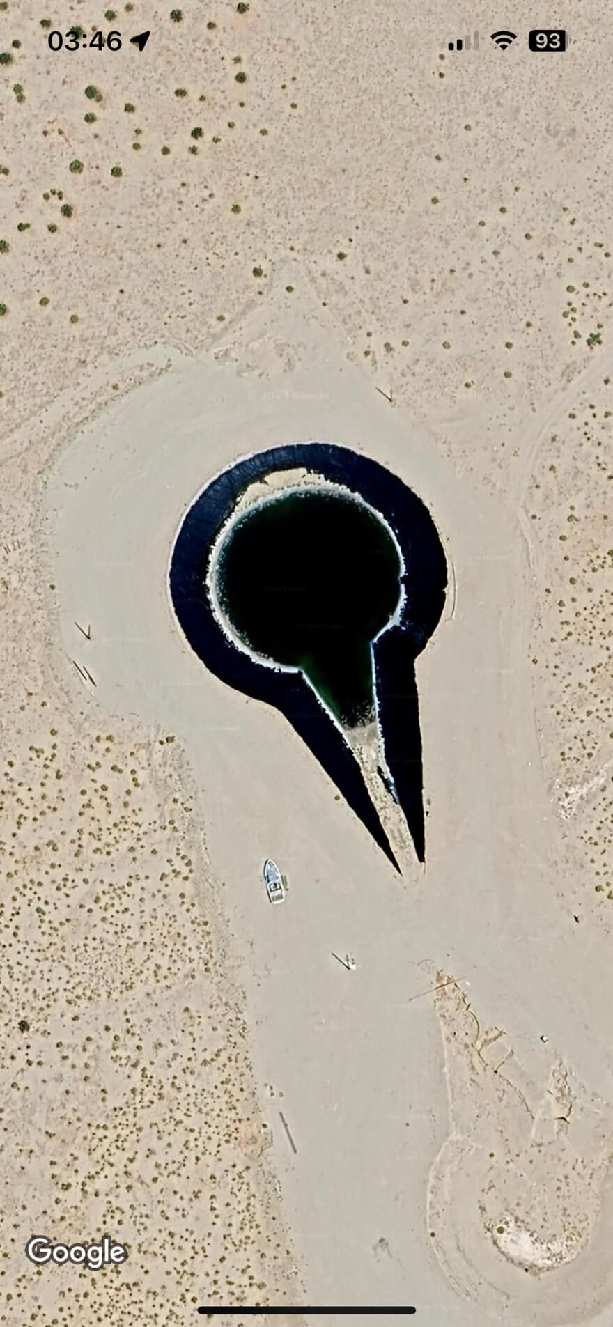 Strange Google Earth finding: a large, dark, drop-shaped pattern in a sandy terrain with nearby scattered vegetation.