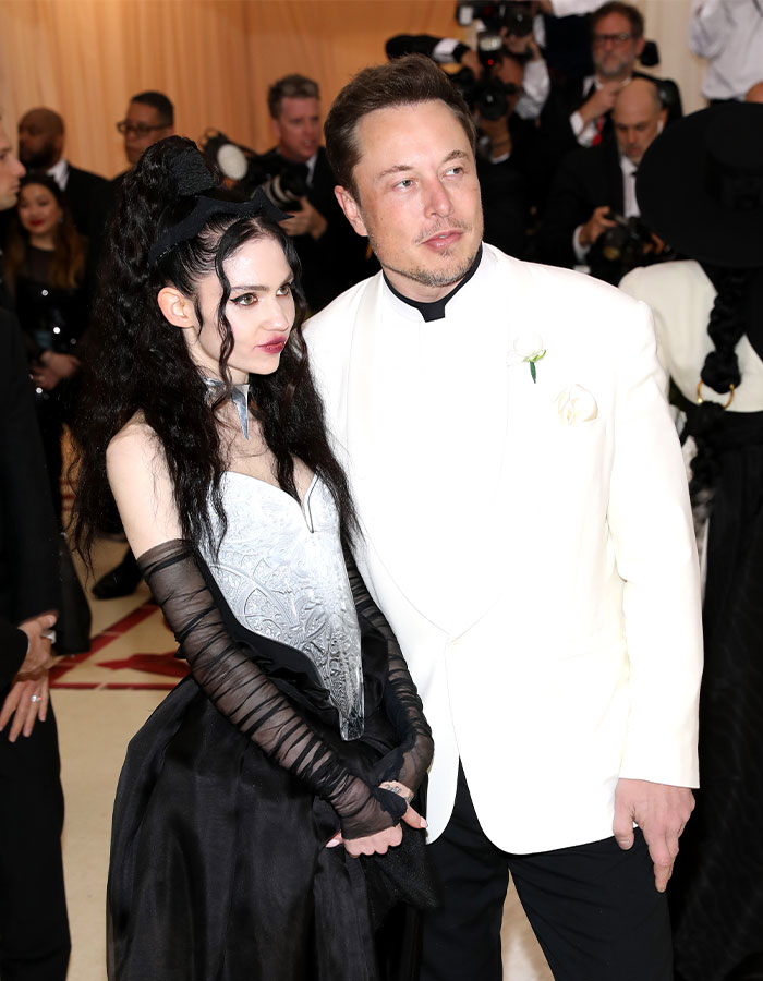 Couple posing at an event, with Grimes responding to controversial salute accusations; Elon Musk in a white tuxedo. Couple posing at an event, with Grimes responding to controversial salute accusations; Elon Musk in a white tuxedo.