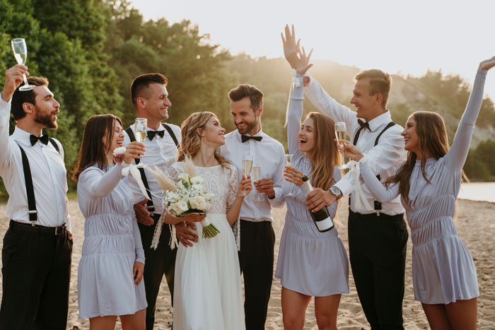 Wedding party celebrating outdoors with champagne, bride holding bouquet, smiling, surrounded by happy guests.