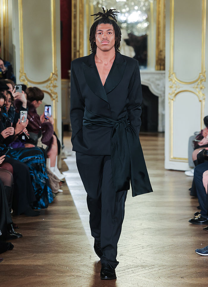 Young male model making runway debut at Paris Fashion Week in a black suit, walking confidently on a wooden floor.