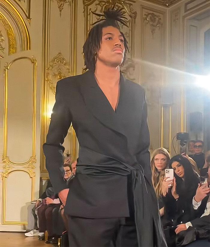 Model walks runway at Paris Fashion Week in sleek black suit, with audience in the background.
