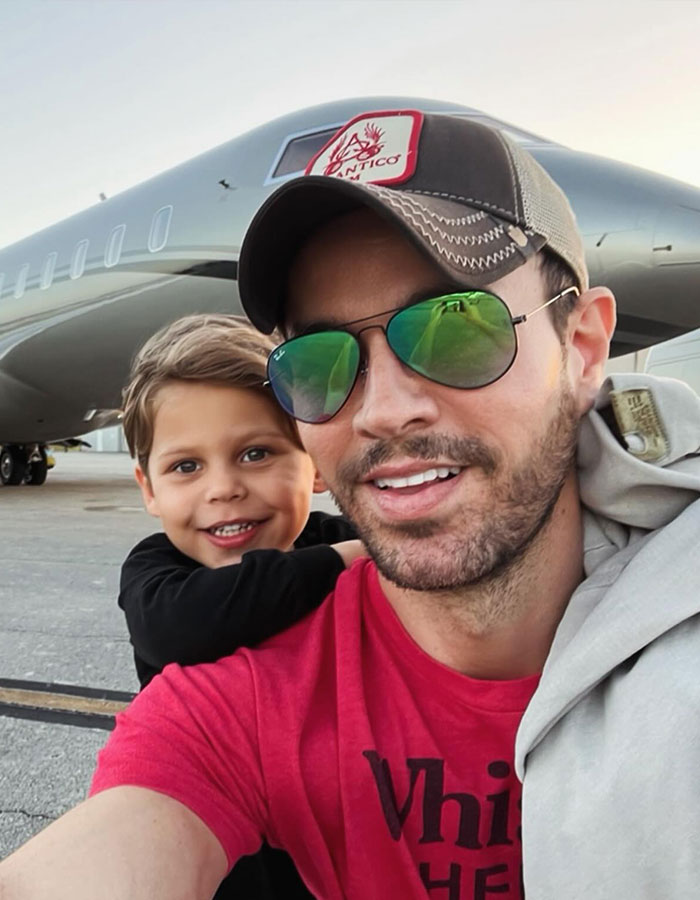 A man in a red shirt and hat with sunglasses smiles with a child, standing in front of a private jet at an airport.