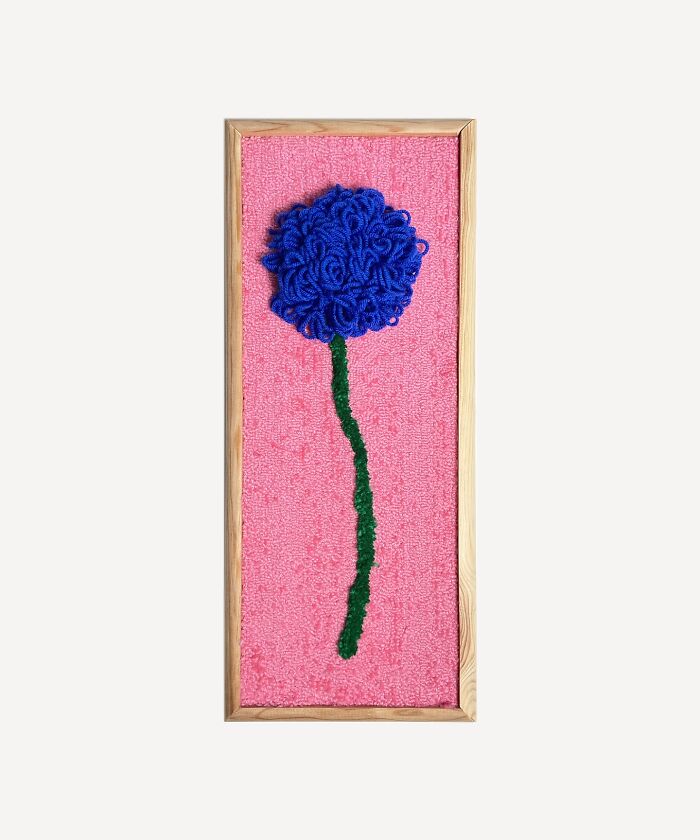 Tufted artwork by Cléa Delogu featuring a blue flower on a pink background in a wooden frame.