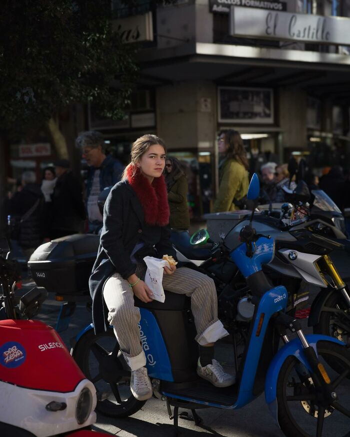 A person sits on a blue scooter, holding a snack, surrounded by bustling street life.