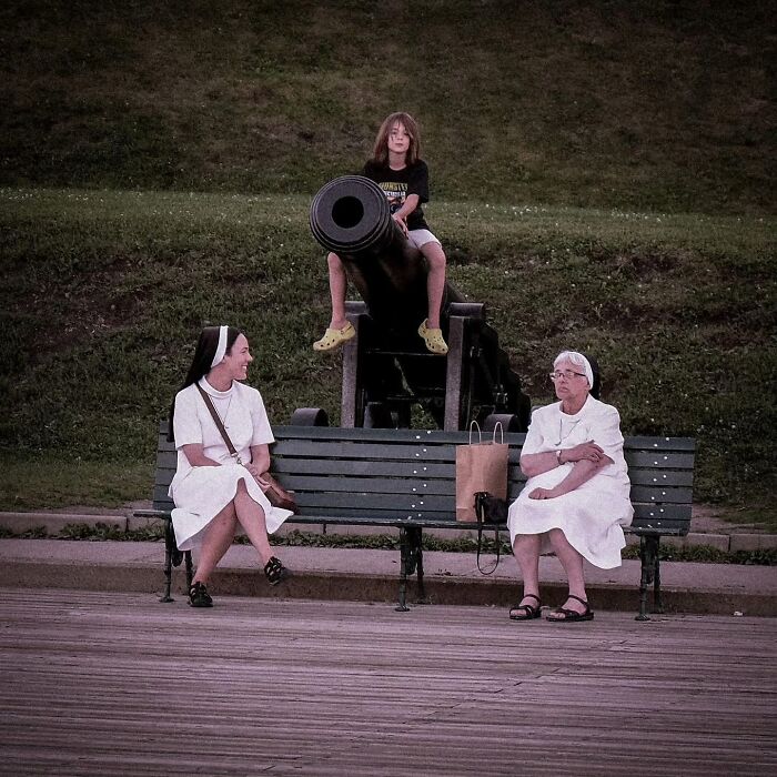 Street photography capturing two women on a bench with a child atop a cannon in the background.