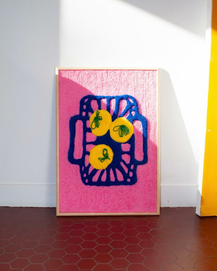 Tufted artwork by Cléa Delogu featuring abstract lemon design on a pink background, framed and leaning against a white wall.