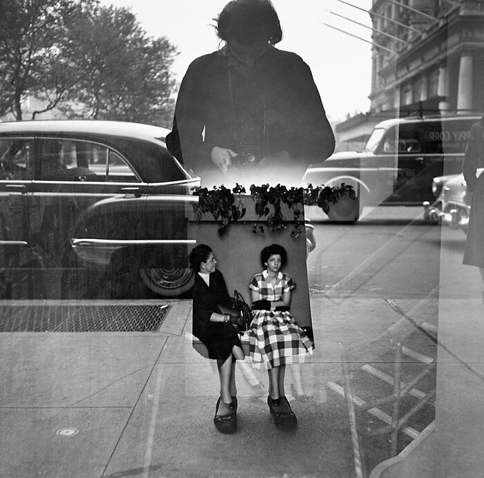 Street photography featuring a reflection of a photographer, vintage car, and two women sitting inside a building.