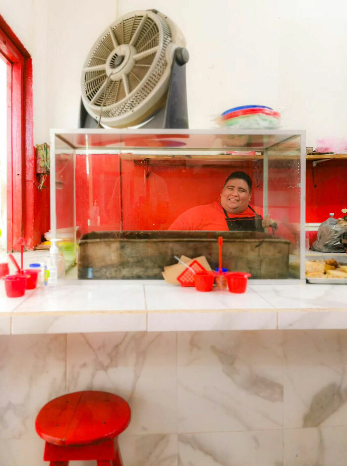 Person in red shirt behind a food counter with fan above, capturing street photography essence.