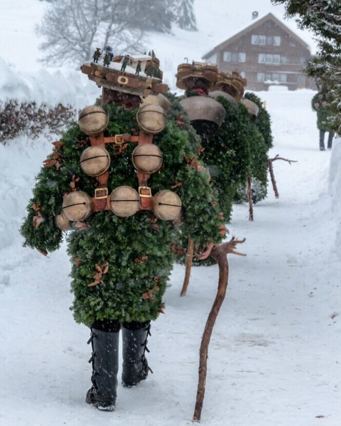 Street photography of festively dressed people in snow, carrying bells and sticks, with a wooden house in the background.