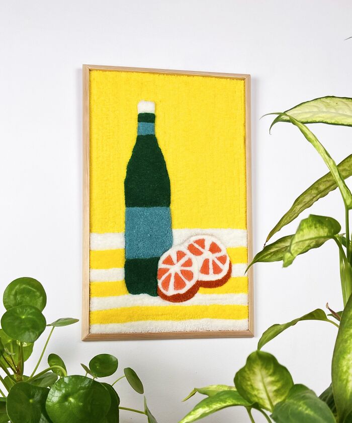 Tufted artwork by Cléa Delogu featuring a bottle and sliced citrus on a vibrant yellow background, surrounded by plants.