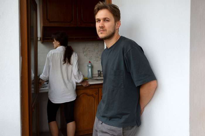 Man standing against a wall, looking away, while a woman is in the kitchen. Man standing against a wall, looking away, while a woman is in the kitchen.