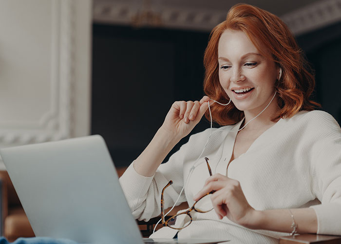 Woman smiling during a video call, embracing uniqueness, wearing a white sweater and headphones, with a laptop.