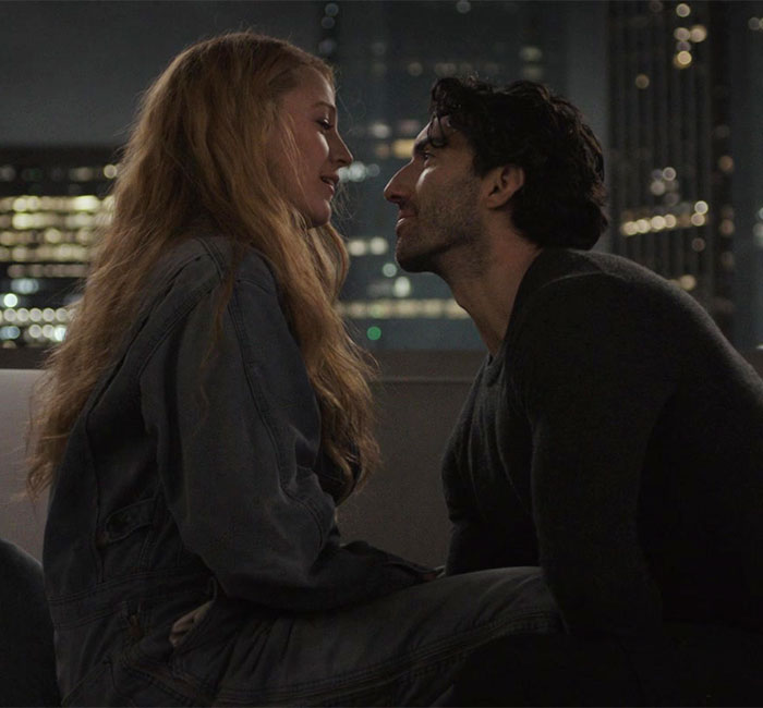 Two people sitting closely on a rooftop at night, city lights in the background.