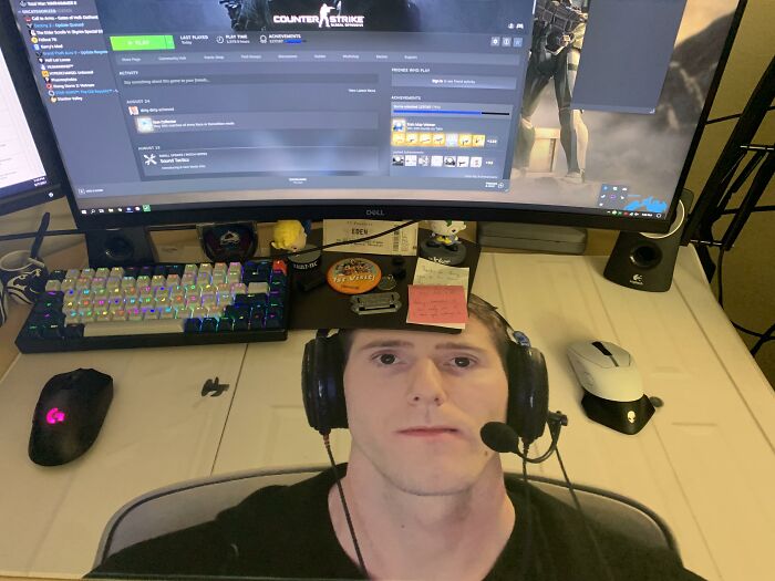 Gamer setup with a large monitor, colorful keyboard, and gaming headset near a humorous cardboard face gift. 