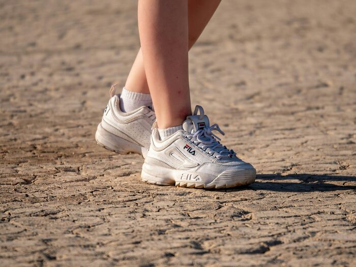 Person wearing white FILA sneakers on a dry, c*****d ground, symbolizing choosing comfort over trends.