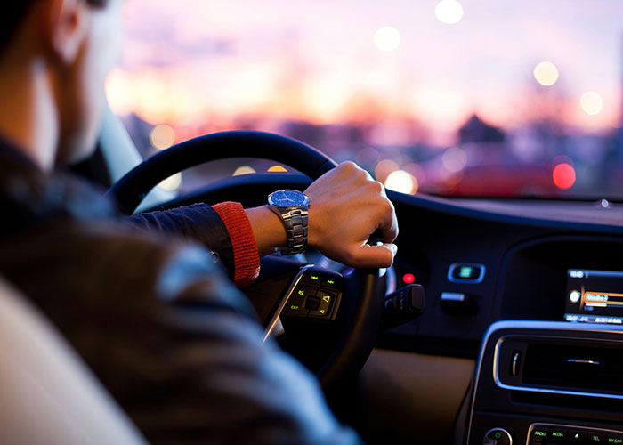 Person driving a car at sunset, wearing a wristwatch, with city lights in the background.