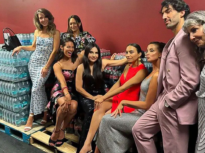 Group posing in a basement setting, Justin Baldoni in a pink suit beside water bottles, surrounded by smiling people.