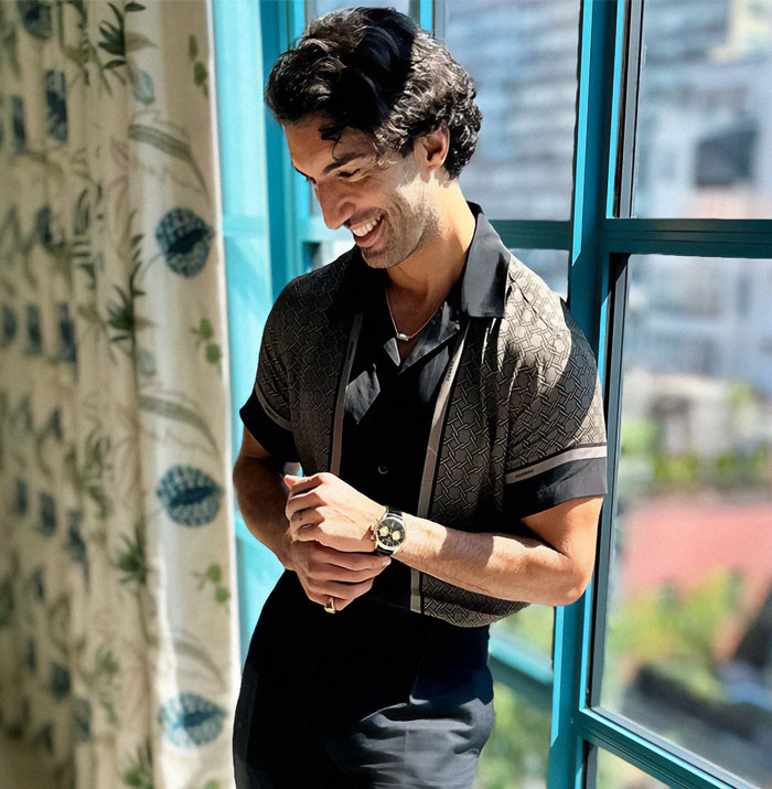 Man in a patterned shirt standing by a window, smiling softly in natural light.
