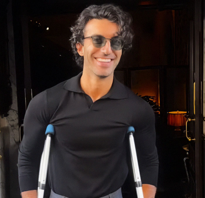 Justin Baldoni in a black shirt and sunglasses, smiling with crutches outside a dimly lit venue; associated with leaked text exchanges.