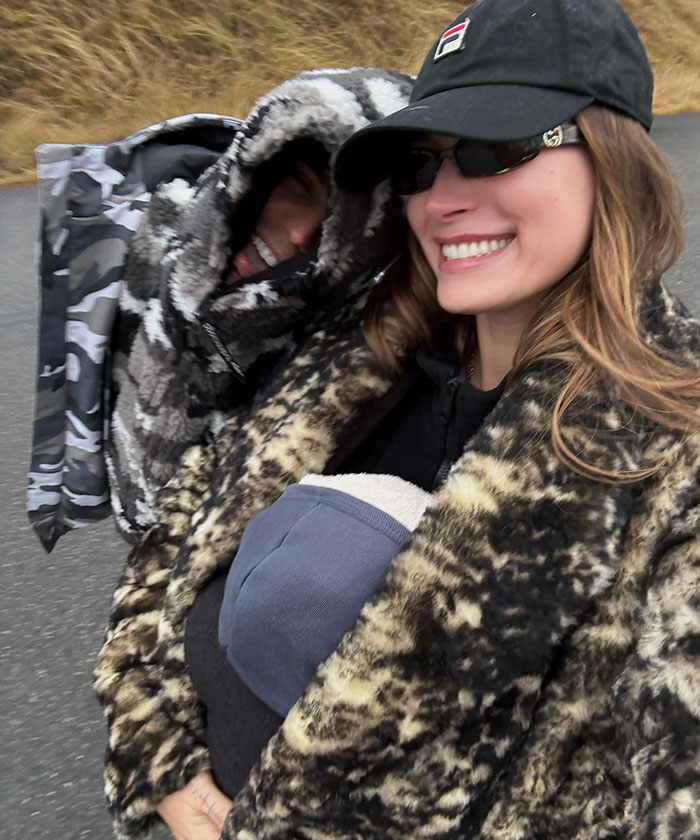Hailey Bieber smiling in a fur coat next to a hooded person on a nature walk. Hailey Bieber smiling in a fur coat next to a hooded person on a nature walk.