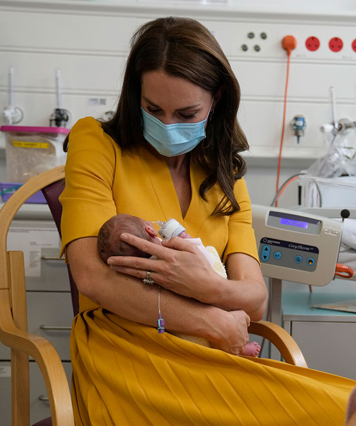 A woman in a yellow dress, wearing a mask, holds a baby in a hospital setting, engagement ring visible.