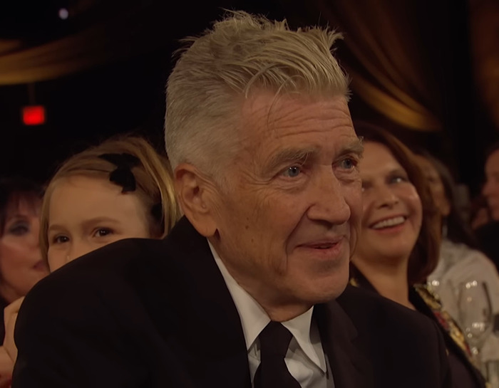David Lynch in a suit smiling in an audience setting, with people seated around him.