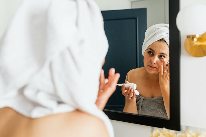 Woman applying skincare product in mirror, wrapped in a towel, highlighting false advertisement in beauty industry.