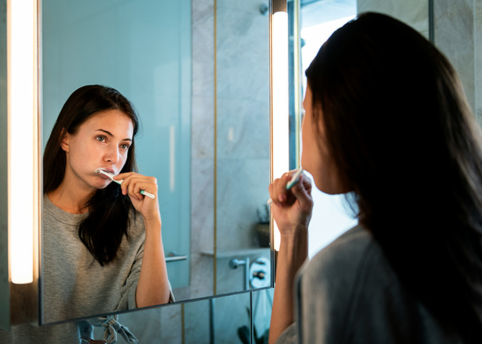 Woman brushing teeth in front of a bathroom mirror promoting health hacks.