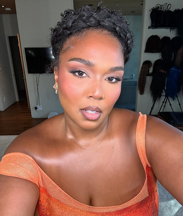 Lizzo flaunting weight loss in an orange top, taking a selfie in a well-lit room. Lizzo flaunting weight loss in an orange top, taking a selfie in a well-lit room.