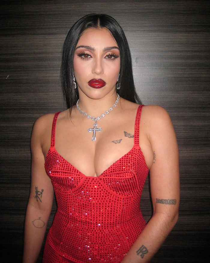 Lourdes Leon in a sparkling red bodysuit with tattoos, standing against a wooden backdrop.