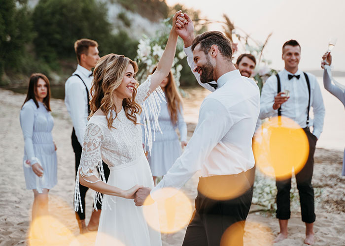 Bride dancing with groom on a beach with friends, smiling despite canceled wedding cake order.