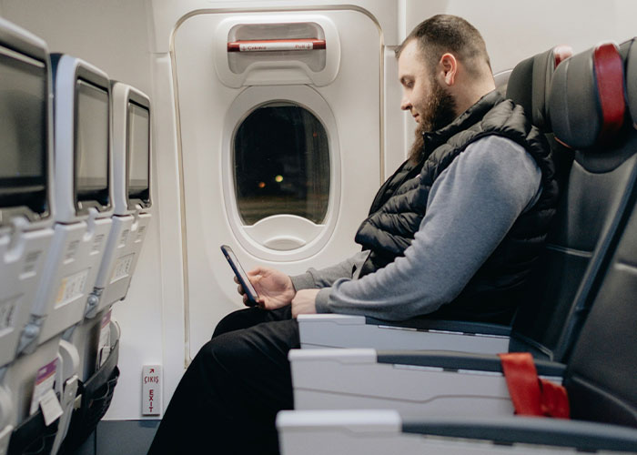 Man in window seat on airplane, wearing gray sweater and black vest, using his phone. Man in window seat on airplane, wearing gray sweater and black vest, using his phone.