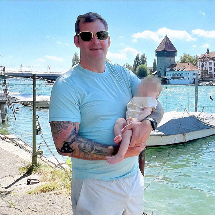 Man with tattoos holding a baby by a lakeside, wearing sunglasses and a blue shirt.