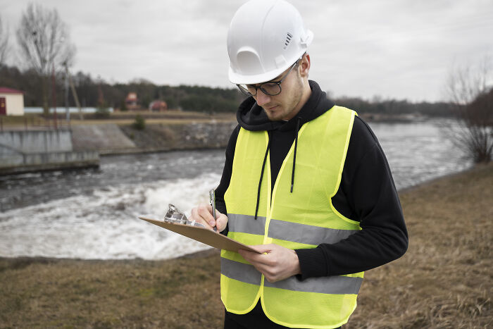 Man in a hard hat and safety vest, reviewing notes by a river, highlighting high-paying remote jobs in engineering.