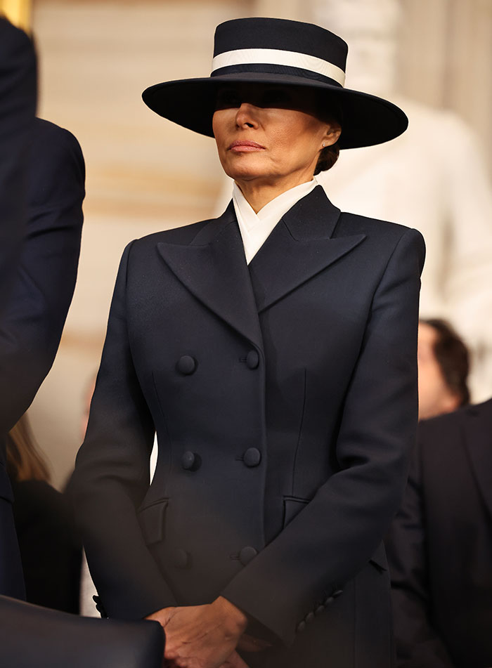 Melania Trump in a dark coat and hat, standing with a neutral expression during a public event. Melania Trump in a dark coat and hat, standing with a neutral expression during a public event.