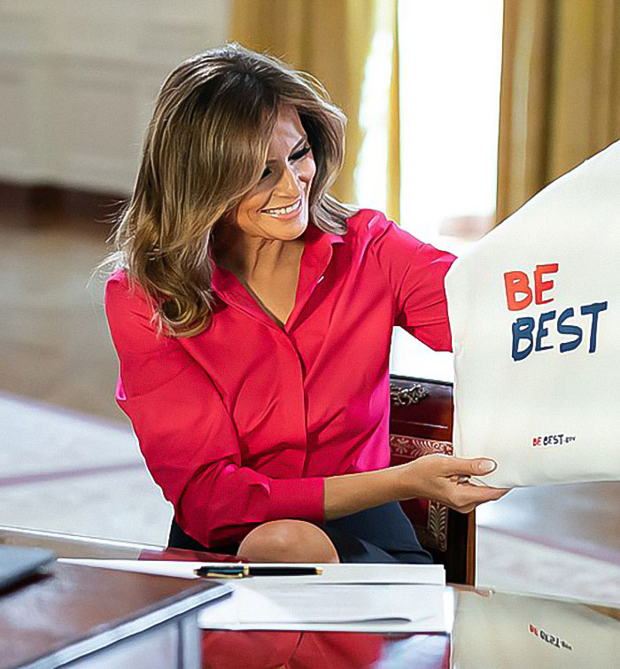 Melania Trump holding "Be Best" material, seated in an elegant room, wearing a bright pink blouse. Melania Trump holding "Be Best" material, seated in an elegant room, wearing a bright pink blouse.