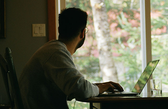 Man in a casual setting, working on a laptop near a window, reflecting on NSFW questions women are too shy to ask.