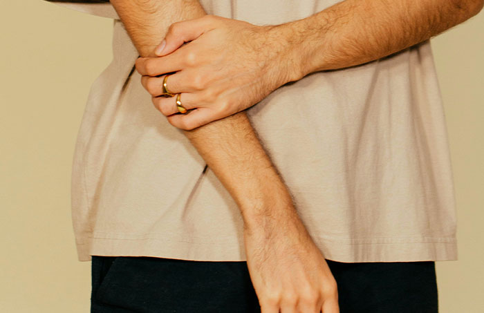 Man in beige shirt holding his arm, wearing rings, addressing questions women are too shy to ask.