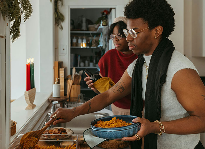 Person serving spicy food in a kitchen, with another person preparing ingredients.