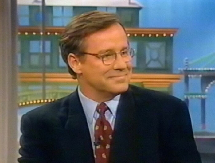 Person in a suit and glasses smiling on a TV show set. Celebrity deaths discussed.