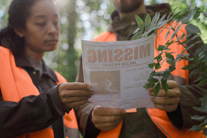 "Two people in safety vests examine a missing person flyer in a forest, highlighting man&rsquo;s mystery disappearance.