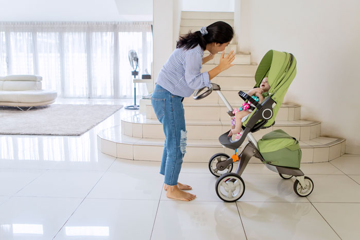 Woman facing a stroller in a modern living area, with sunlight streaming in through large windows.