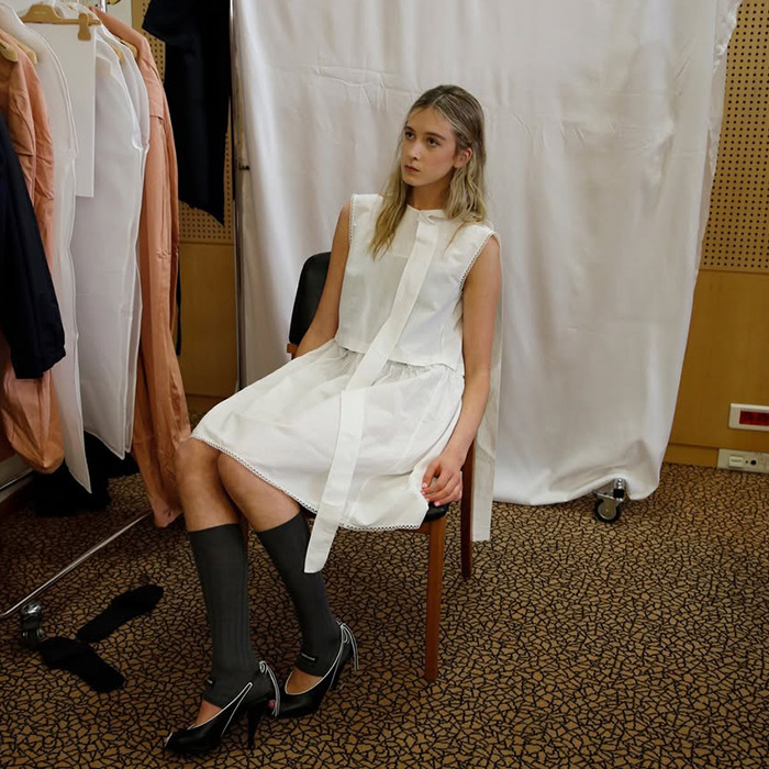 Sunday Rose in white dress and black heels sitting, backstage at a fashion show.