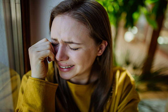 A woman in a yellow top, visibly upset and crying, sits by a window. A woman in a yellow top, visibly upset and crying, sits by a window.