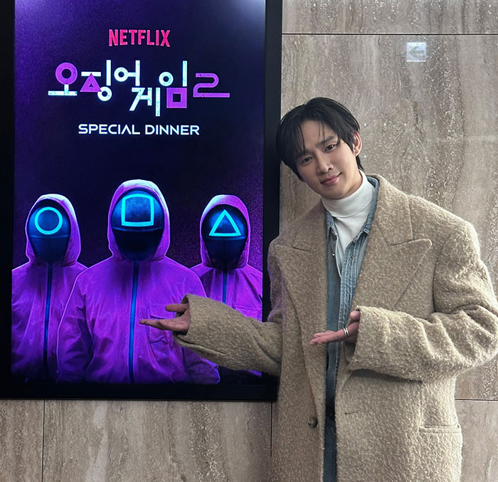 Actor with a poster for Squid Game special dinner event, wearing a beige coat and posing indoors. Actor with a poster for Squid Game special dinner event, wearing a beige coat and posing indoors.