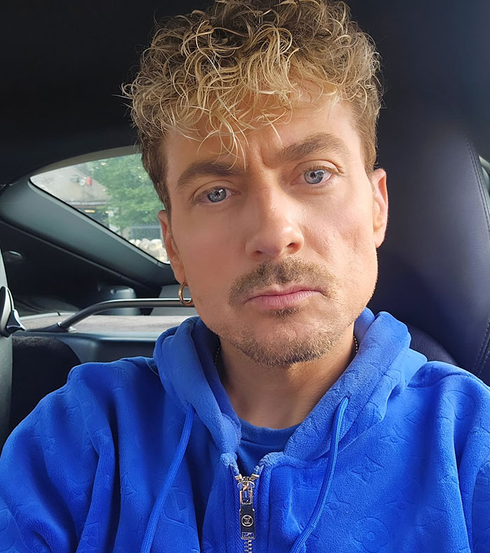 Man in a car wearing a blue hoodie with curly hair and an earring.
