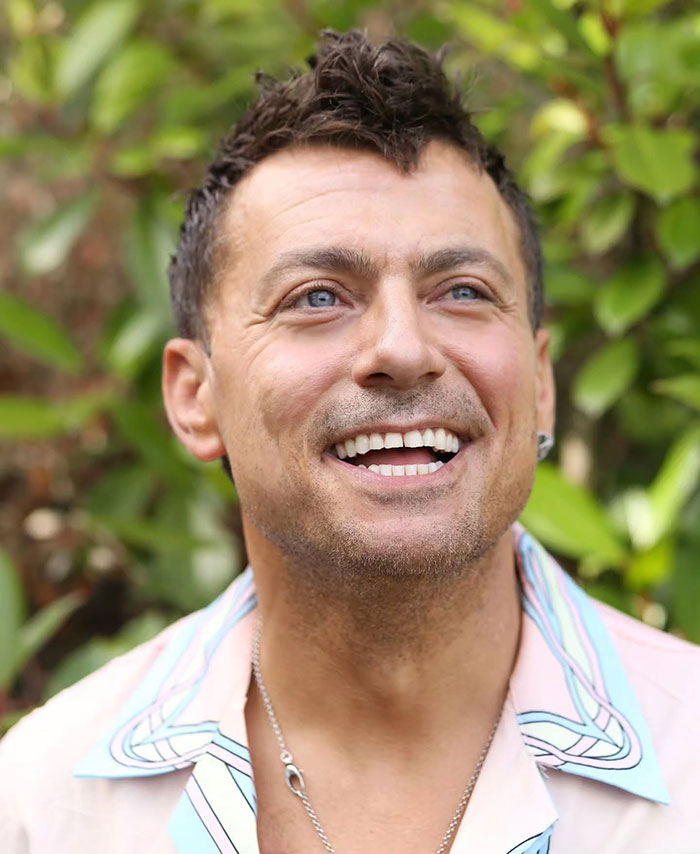 Man smiling outdoors, wearing a light shirt, with green foliage in the background.