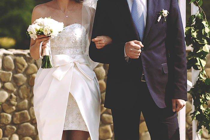 Bride holding flowers, in a wedding dress with an escort in a suit, walking down an outdoor aisle.