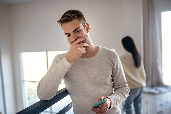 Man in gray sweater looking contemplative with smartphone, woman in background, related to attention-seeking SIL in wedding context.