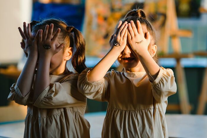 Twin girls covering their eyes with hands, painted with eyes, in soft lighting, embodying weird unsolved mysteries.